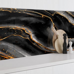 Puede incluir: Salpicadero de cocina abstracto negro y dorado con un patrón de mármol. El salpicadero está detrás de una encimera blanca con tablas de cortar de madera y saleros y pimenteros.