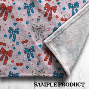 May include: A light pink blanket with a repeating pattern of red, white, and blue bows, stars, cherries, and popsicles. The blanket is folded over, revealing a soft, white interior. The words "SAMPLE PRODUCT" are printed at the bottom.