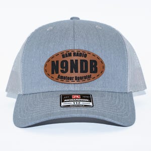 May include: A gray trucker hat with a mesh back and a curved brim. The hat features a brown oval patch with the text "HAM RADIO N9NDB Amateur Operator". A small black and white tag is visible on the front.