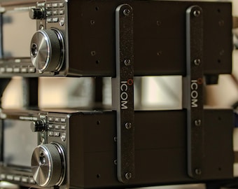 Könnte beinhalten: Zwei schwarze ICOM Funktransceiver, die auf einem Regal gestapelt sind und von schwarzen Halterungen mit dem ICOM-Logo gehalten werden. Jedes Funkgerät hat einen großen silbernen Knopf und ein Display mit Tasten.