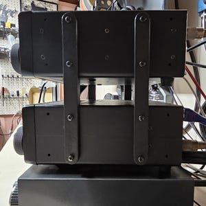 May include: Two black electronic devices stacked on top of each other. The devices are mounted on a black metal stand with brackets. The devices have a knob on the left side and a series of holes on the front.