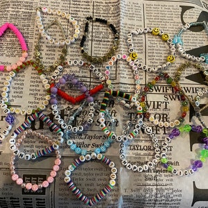 May include: A collection of colorful beaded bracelets with various words and designs, including "Taylor Swift", "Lavender Haze", "1989", "Our Song", "Happiness", and "Willow".