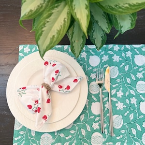 May include: A white dinner plate with a white napkin folded into a bow with red berries printed on it. The plate is set on a green placemat with a white pomegranate pattern.
