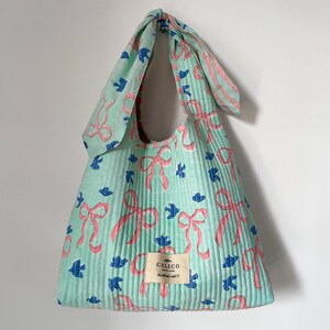 May include: A mint green tote bag with a repeating pattern of pink bows and blue bird shapes. The bag has a knotted handle and a small tag that reads "CALICO". The bag is made of a ribbed fabric.