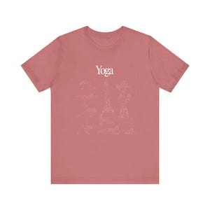 May include: Dusty rose t-shirt with the word "Yoga" in white script at the top. Below the word are several line drawings of yoga poses in white. The shirt is made of a soft material.