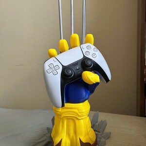 May include: A yellow and blue hand sculpture with three long silver claws extending upward, holding a white and black video game controller. The base of the sculpture is yellow with brown accents and features gray jagged rock-like details.