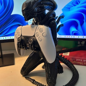 May include: A black Xenomorph figurine from the Alien franchise, sculpted in a crouching position with its hands extended forward to hold a white and black wireless video game controller. The figure features a smooth, elongated head and tail.
