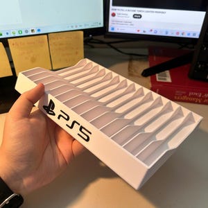 Puede incluir: Un soporte de almacenamiento de discos de juego de plástico blanco con una serie de ranuras verticales para organizar varios discos. El lateral del soporte muestra el logotipo negro de PlayStation 5 y el texto PS5 impreso en la superficie.