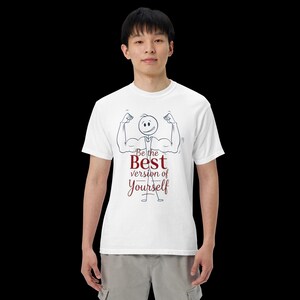 May include: White t-shirt featuring a stick figure flexing its muscles, with the text "Be the Best version of Yourself" in a handwritten style. The design is in black and burgundy. The shirt is worn by a person.