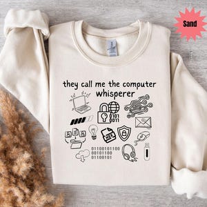 Peut inclure: Un sweat-shirt couleur sable avec l'inscription "they call me the computer whisperer" et des icônes informatiques noires. Le design comprend un ordinateur portable, un cadenas, un circuit imprimé et du code binaire.