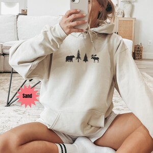 May include: A sand-colored hoodie with a front pocket and a drawstring hood. The hoodie features a black graphic of a bear, two trees, and a moose. The word "Sand" is in a pink starburst graphic.
