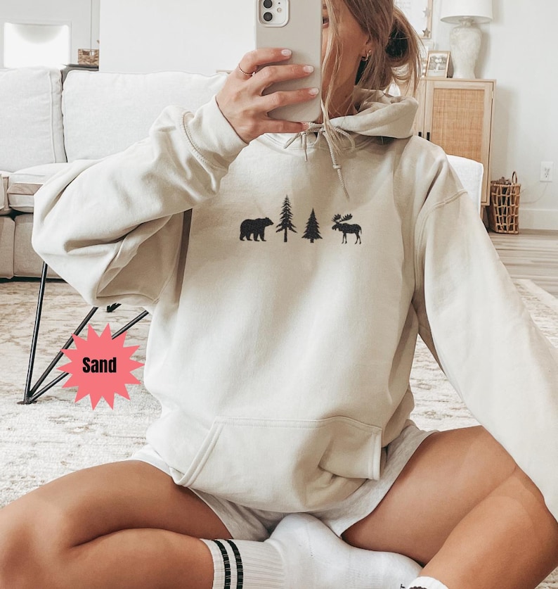 May include: A sand-colored hoodie with a front pocket and a drawstring hood. The hoodie features a black graphic of a bear, two trees, and a moose. The word "Sand" is in a pink starburst graphic.