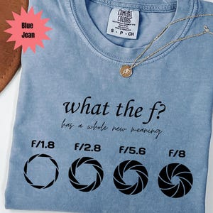 May include: A blue jean coloured t-shirt with the text "what the f? has a whole new meaning" and f-stop aperture graphics. A gold necklace with a pendant is draped across the shirt. The shirt has a Comfort Colors tag.