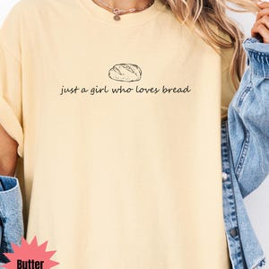 May include: Butter-colored t-shirt with a black illustration of a loaf of bread and the text "just a girl who loves bread." The shirt has a crew neck and short sleeves. The model is wearing a denim jacket.