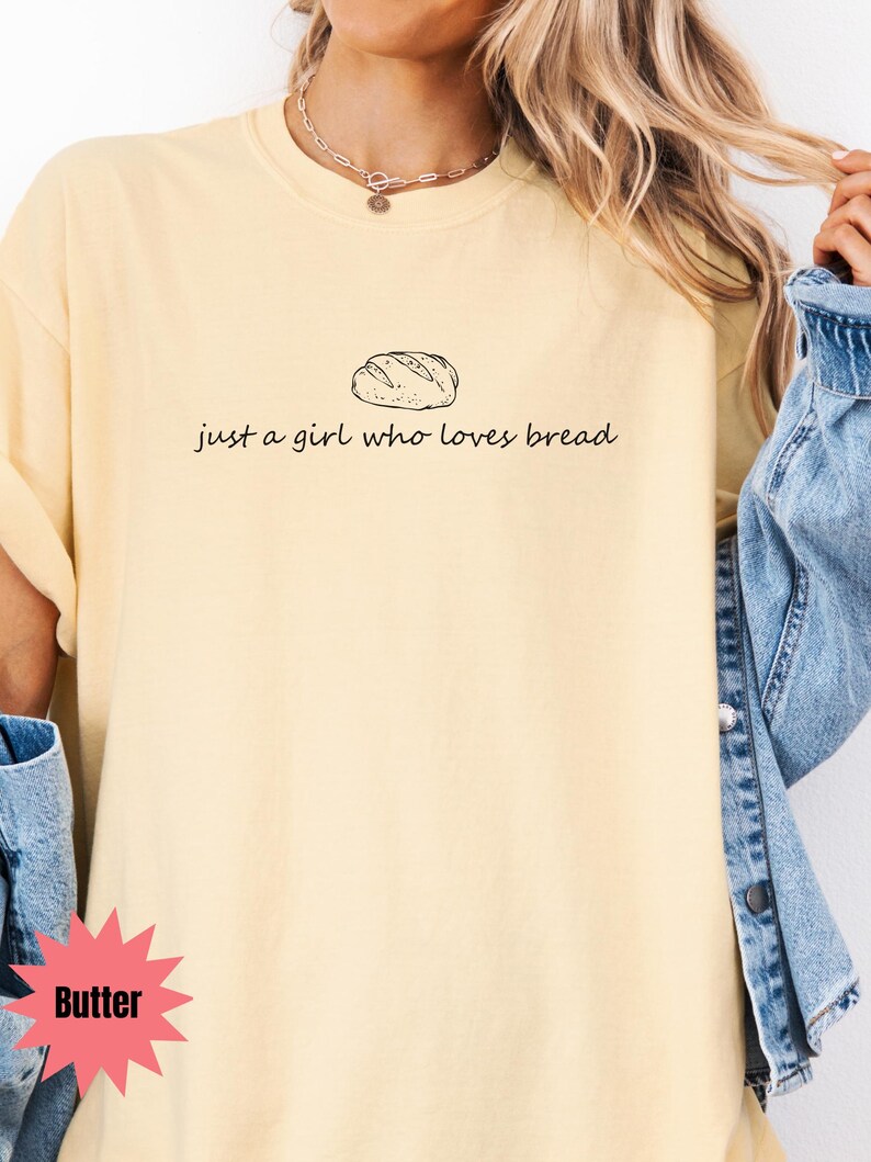 May include: Butter-colored t-shirt with a black illustration of a loaf of bread and the text "just a girl who loves bread." The shirt has a crew neck and short sleeves. The model is wearing a denim jacket.