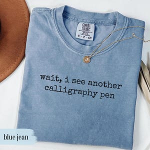 May include: A blue jean colored t-shirt with the text "wait, i see another calligraphy pen" in black. A gold necklace with a pendant is draped across the shirt. The shirt has a crew neck and short sleeves.