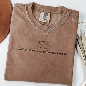 May include: A brown Comfort Colors t-shirt with the text "just a girl who loves bread" and a bread loaf graphic. A silver necklace with a pendant is on the shirt.