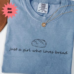 May include: Blue jean Comfort Colors t-shirt with the text "just a girl who loves bread" and a bread loaf graphic. A gold necklace with a round pendant is on the shirt.