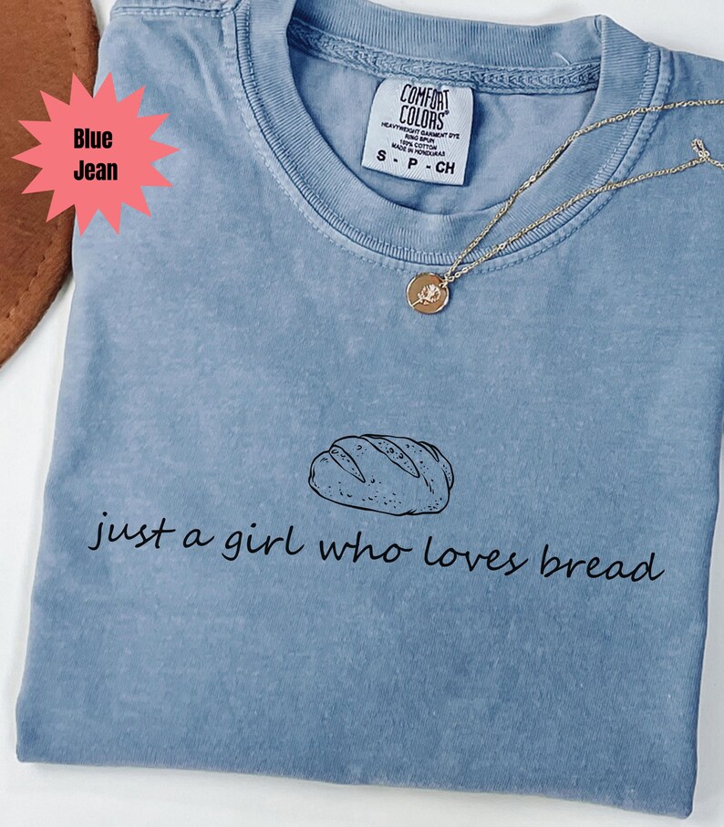 May include: Blue jean Comfort Colors t-shirt with the text "just a girl who loves bread" and a bread loaf graphic. A gold necklace with a round pendant is on the shirt.