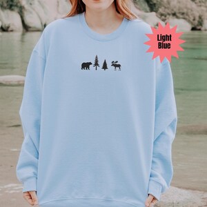 May include: Light blue sweatshirt with a black bear, two trees, and a moose graphic. The text "Light Blue" is in a pink starburst. The sweatshirt is a solid light blue color.