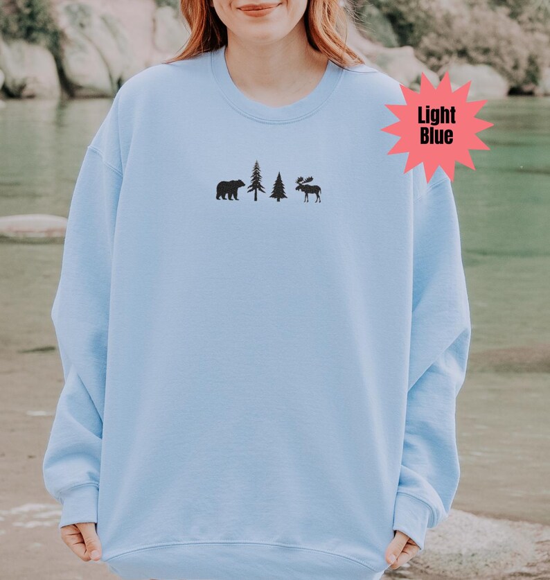 May include: Light blue sweatshirt with a black bear, two trees, and a moose graphic. The text "Light Blue" is in a pink starburst. The sweatshirt is a solid light blue color.