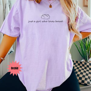 May include: Orchid-colored t-shirt with the text "just a girl who loves bread" and a bread loaf graphic. The shirt is a light purple color and has short sleeves. The text and graphic are in black.
