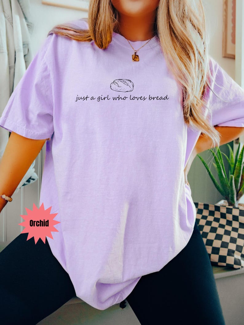 May include: Orchid-colored t-shirt with the text "just a girl who loves bread" and a bread loaf graphic. The shirt is a light purple color and has short sleeves. The text and graphic are in black.