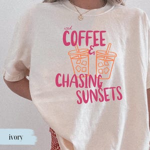 May include: Cream-coloured t-shirt featuring the text "iced COFFEE & CHASING SUNSETS" in pink and orange. The design includes two iced coffee cups. A casual wear item.