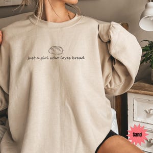 May include: Sand-colored sweatshirt with the text "just a girl who loves bread" and a bread loaf illustration. The long-sleeved top is a neutral color and has a relaxed fit. The text and illustration are in black.