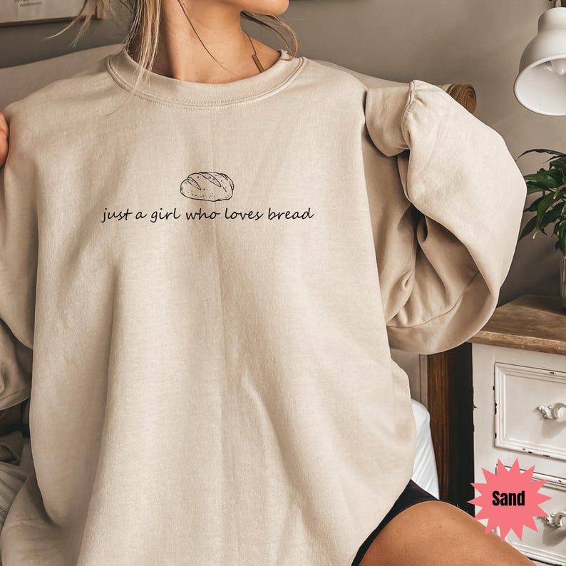 May include: Sand-colored sweatshirt with the text "just a girl who loves bread" and a bread loaf illustration. The long-sleeved top is a neutral color and has a relaxed fit. The text and illustration are in black.