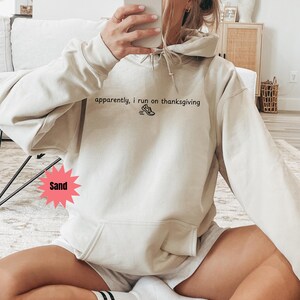 May include: Sand-colored hoodie with the text "apparently, I run on thanksgiving" and a graphic of a running shoe. The hoodie has a front pocket and a drawstring hood. The model is wearing shorts.