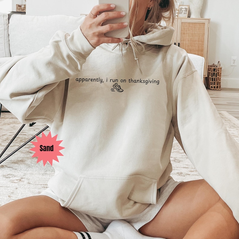 May include: Sand-colored hoodie with the text "apparently, I run on thanksgiving" and a graphic of a running shoe. The hoodie has a front pocket and a drawstring hood. The model is wearing shorts.
