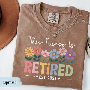 May include: Espresso-colored t-shirt with the text "This Nurse Is Retired EST. 2026" in white and colorful letters. The design includes a row of flowers in yellow, pink, blue, and purple. A silver necklace with a small pendant is on the shirt.
