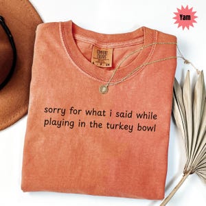 May include: A coral-colored t-shirt with the text "sorry for what I said while playing in the turkey bowl." A gold necklace with a pendant rests on the shirt. The shirt has a Comfort Colors tag.