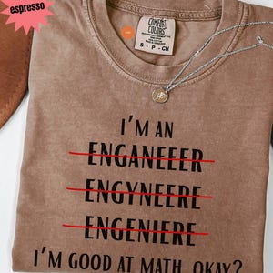 May include: Espresso-coloured t-shirt with the text "I'M AN ENGANEER, ENGYNEERE, ENGENIERE, I'M GOOD AT MATH, OKAY?" crossed out in red. A silver necklace with a pendant is on the shirt.