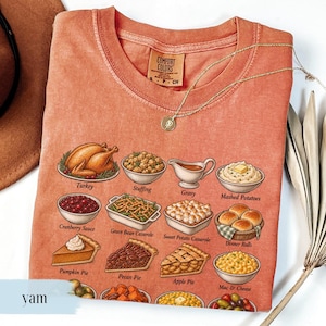 May include: A terracotta-colored short-sleeve t-shirt featuring a graphic print of various Thanksgiving dishes including turkey, stuffing, gravy, mashed potatoes, cranberry sauce, green bean casserole, sweet potato casserole, dinner rolls, and pies.