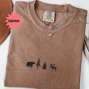 May include: Espresso-colored t-shirt with a crew neck and a small graphic of a bear, trees, and a moose. The shirt has a Comfort Colors tag and a silver necklace with a pendant.