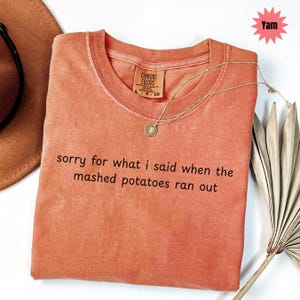 May include: A coral-colored Comfort Colors t-shirt with the text "sorry for what I said when the mashed potatoes ran out." A gold necklace with a pendant rests on the shirt. A brown hat and decorative leaf are in the background.