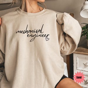 May include: Sand-colored sweatshirt with the words "mechanical engineer" in a cursive font, and a small heart symbol. The sweatshirt has a crew neck and long sleeves.