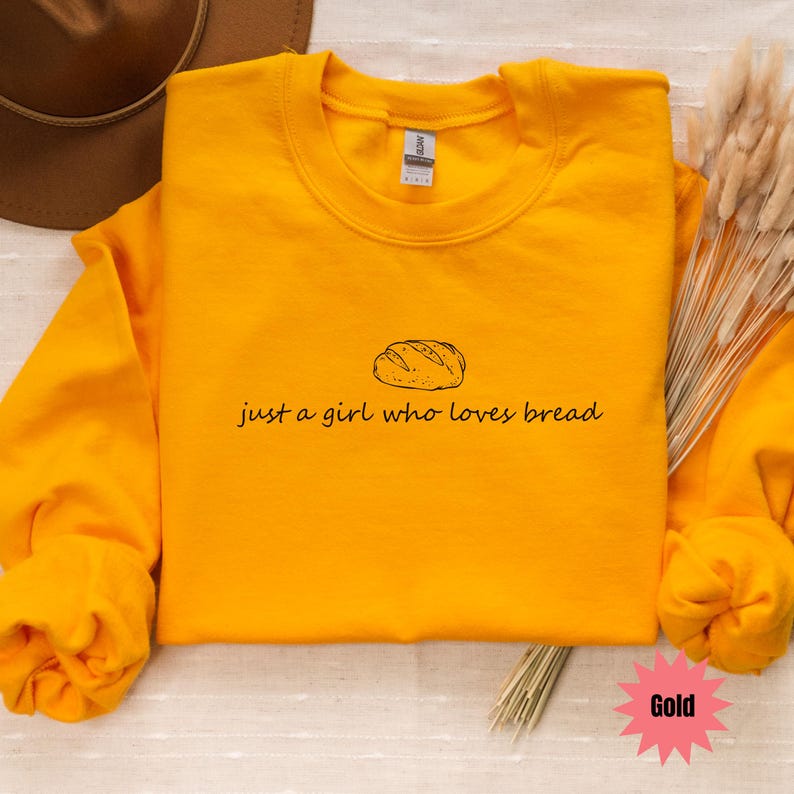 May include: A gold-colored sweatshirt with the phrase "just a girl who loves bread" and a bread illustration. The sweatshirt is a crew neck style and has long sleeves. The word "Gold" is in a pink starburst.