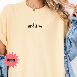 May include: Butter-colored t-shirt with a small black graphic of a bear, two trees, and a moose. The word "Butter" is in a pink starburst shape. The shirt has a relaxed fit and short sleeves.