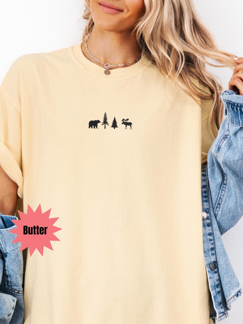 May include: Butter-colored t-shirt with a small black graphic of a bear, two trees, and a moose. The word "Butter" is in a pink starburst shape. The shirt has a relaxed fit and short sleeves.