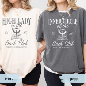 May include: Two oversized t-shirts, one ivory and one pepper, with the text "HIGH LADY of the Bach Club" and "INNER CIRCLE of the Bach Club". Each shirt has a graphic design with a book and a stylized illustration. The text "Sophia's Bachelorette" is also included.