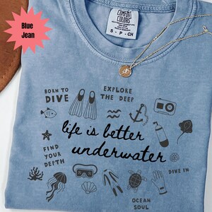 May include: Blue jean Comfort Colors t-shirt with a gold necklace. The shirt features black illustrations of diving equipment and the phrase "life is better underwater". Additional text includes "Born to Dive" and "Explore the Deep".