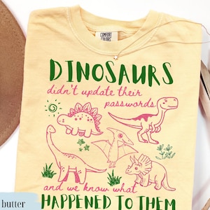 May include: A pale yellow t-shirt featuring a graphic print with green and pink text that reads: DINOSAURS didn't update their passwords and we know what HAPPENED TO THEM. The design includes line-art illustrations of various dinosaurs.