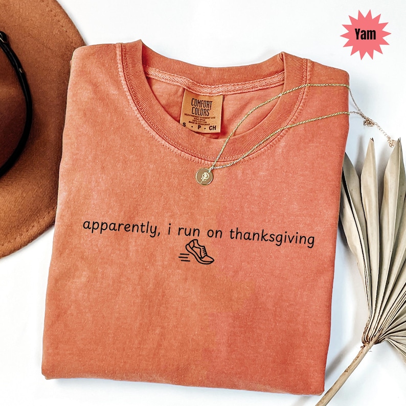 May include: A coral-colored t-shirt with the text "apparently, i run on thanksgiving" and a running shoe graphic. A gold necklace with a pendant rests on the shirt. The shirt is labeled "Comfort Colors".