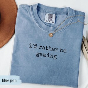 May include: A blue jean colored t-shirt with the phrase "i'd rather be gaming" printed in black. A gold necklace with a pendant is draped across the shirt. The shirt has a crew neck and a tag that reads "Comfort Colors".