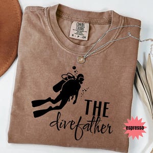 May include: A brown Comfort Colors t-shirt with a black graphic of a scuba diver and the words "THE divefather". A silver necklace with a pendant is on the shirt. The word "espresso" is in a pink starburst.