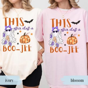May include: Two short-sleeve t-shirts in ivory and blossom pink. The graphic features a ghost wearing a purple bow, heart-shaped sunglasses, and trainers, holding a drink. Text reads: THIS office staff is BOO-JEE, accompanied by a pumpkin and bat.