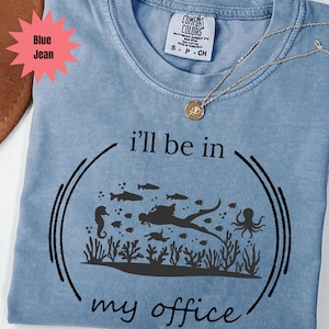 May include: A blue jean Comfort Colors t-shirt with the phrase "i'll be in my office" and a scuba diving graphic. The graphic features a diver, fish, seahorse, and an octopus. A gold necklace with a pendant is also visible.